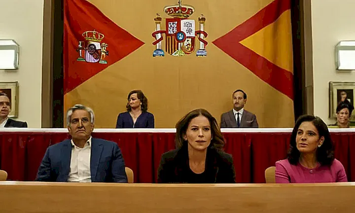 La imagen muestra una reunión de personas en un ambiente acogedor y formal, con un telón de fondo de la bandera española, rodeadas por imágenes geográficas y personas sentadas o de pie con expresiones variadas