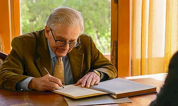 Una persona vestida como funcionario analiza un viejo documento en su escritorio mientras la luz del atardecer de Madrid entra por la ventana detrás de él