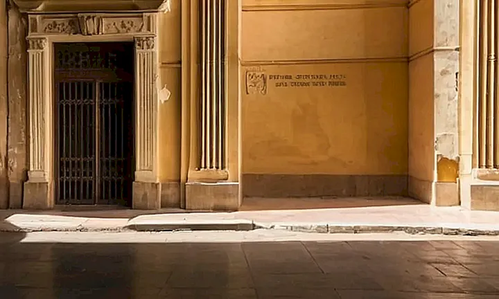 La fachada de un edificio gubernamental español deteriorado es visitado por alguien que camina lentamente, rodeado de un entorno urbano con elementos abandonados y desatendidos