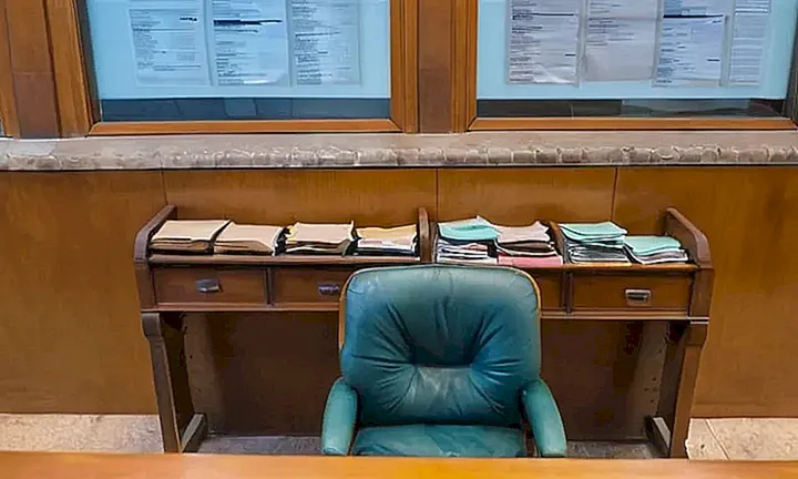 Un oficial con expresión cansada y un escritorio desorganizado trabaja en una oficina descuidada bajo luces fluorescentes, mientras una familia espera su turno en el fondo