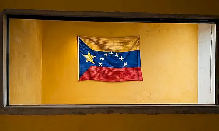 Una bandera venezolana colgando en una pared amarilla con un espacio traslúcido enfocado por una ventana iluminada