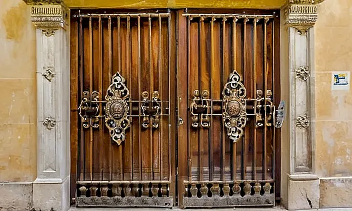 Un edificio antiguo con paredes de estuco gris y balcones blancos está rodeado por un muro de piedra desgastada y una entrada oscura conrejada