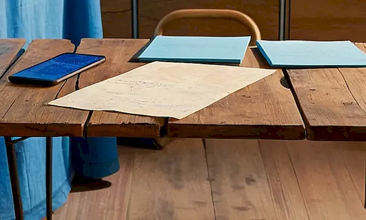 Una mesa de madera gastada con papeles llenos de notas y un teléfono cerca alumbra su superficie desgastada