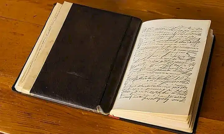 Un libro viejo con una cubierta gris y letras cúficas en la portada descansa sobre un escritorio, rodeado de notas manuscritas y sombreado por el sol