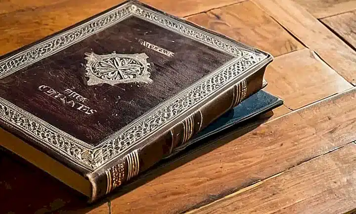 Un libro de cuero antiguas descansa a caballo entre libros, iluminado por una luz suave que destaca los detalles del suelo de madera