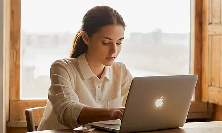 Una joven mujer con el cabello moreno se encuentra sentada enfocándose en leer y escribir en su laptop