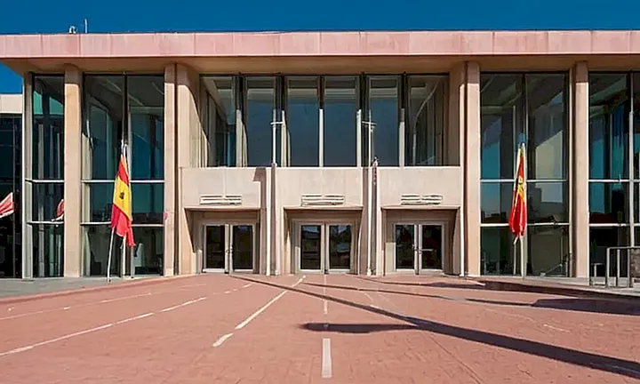 Un edificio gubernamental grisáceo con entrada de vidrio rodeada por un anillo crema y banderas españolas ondeando al viento