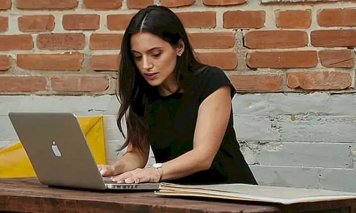 Una mujer está sentada cerca de una mesa de madera y mira su laptop mientras la luz del sol entra por las grietas en un muro de ladrillos