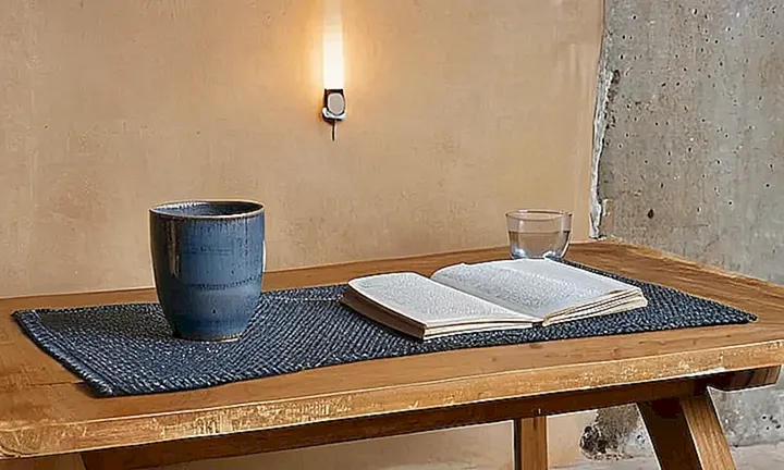 Una mesa de madera descansa frente a una pared gris, junto a objetos como un laptop, papeles anotados y vasijas de cerámica