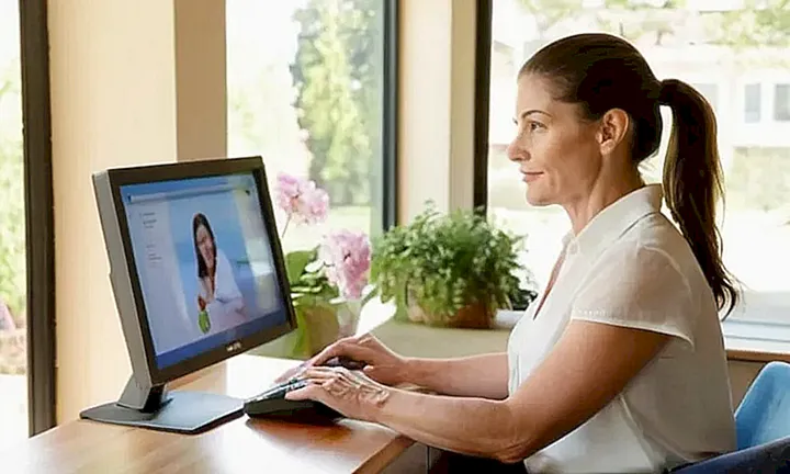 Una mujer de mediana edad con un rostro amable y un ático tranquilo está sentada frente a una computadora, rodeada de fotos personales en una habitación cómoda