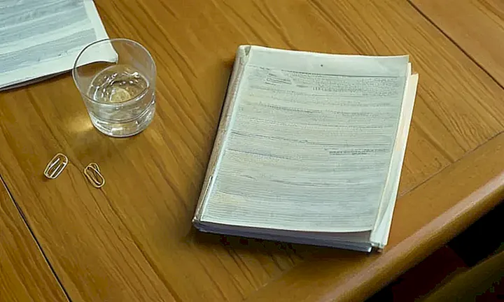 La imagen muestra un espacio con una mesa llena de documentos y un individuo sentado con las manos cruzadas