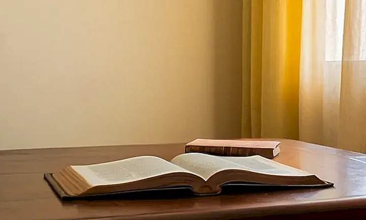 En la habitación hay una mesa antigua con un libro abierto, iluminado por el sol que entra a través de las cortinas amarillas