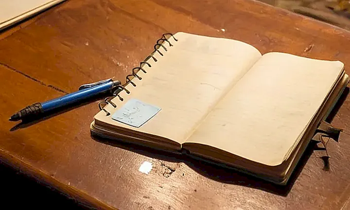 Un escritorio antiguo y desordenado contiene un cuaderno abierto con notas echarpetadas y un bolígrafo gastado