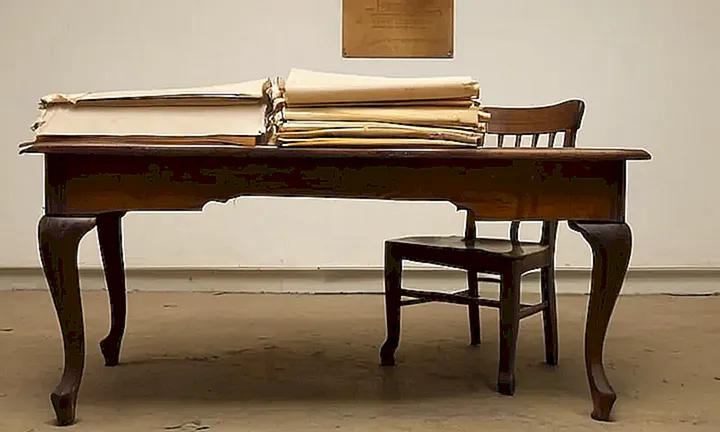 Un espacio de archivo antiguo muestra un tablero desgastado, un silla vieja y un escudo español en forma de plaquita
