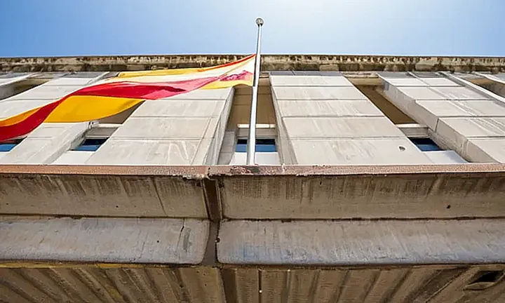 La fachada de un edificio estatal muestra un color cenizo gastado y pintura pelada, con barandillas metálicas oxidadas que destacan junto a una bandera española desvanecida