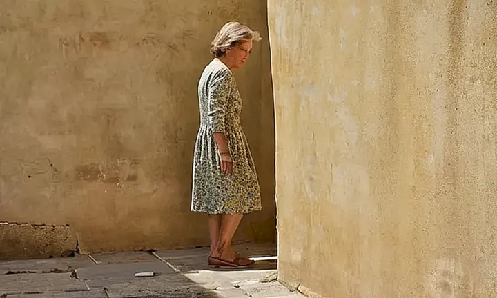 Una mujer madura de expresión cansada permanece de pie en la calle, observando un pequeño modelo de una ciudad antigua bajo el sol inmisericorde del mediodía