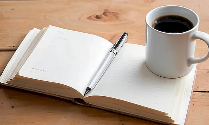 Un libro abierto y una taza de café se encuentran junto a un bolígrafo en una mesa antigua
