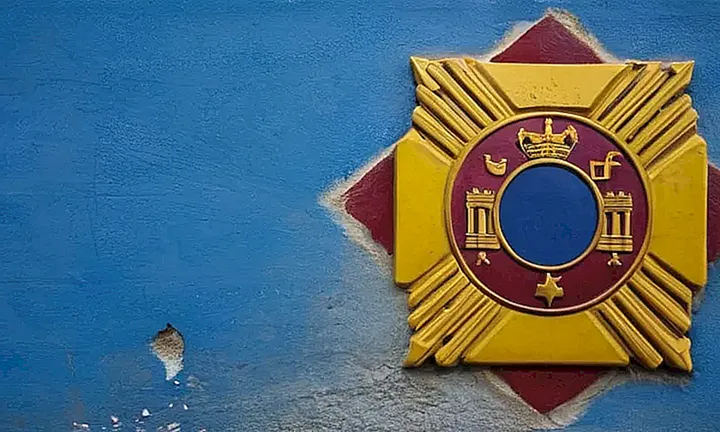 En el fondo hay una pared de concreto gris y desgastado rodeando un logotipo del gobierno español en colores brillantes