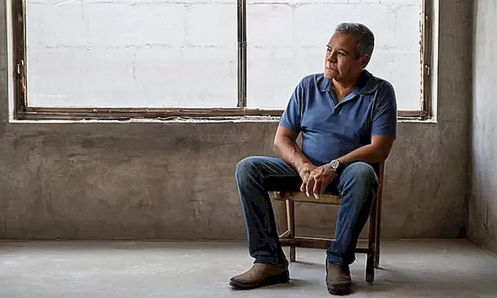Un hombre de mediana edad latinx se sienta en una silla de madera ajada, rodeado por la quietud y un suave brillo de luz solar