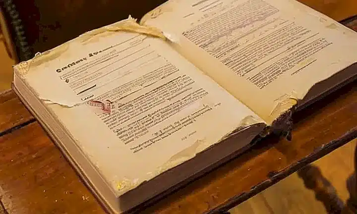 Un antiguo libro de examen de ciudadanía está abierto sobre una mesa roída, con páginas amarillentas y bordes doblados