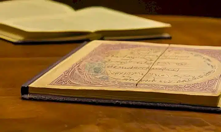 Sobre un escritorio desordenado reposa un libro gastado cubierto de cuero marrón, con páginas amarilleadas y textos en pequeña letra decorados con caligrafía