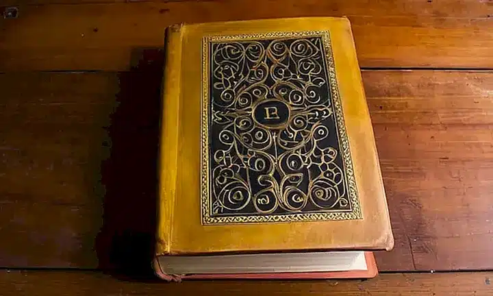 Un libro antiguo de cuero desgastado descansa entreabrído sobre una mesa de madera, mostrando sucinta su tapa con el nombre del autor Carlos Poveda en letras negras