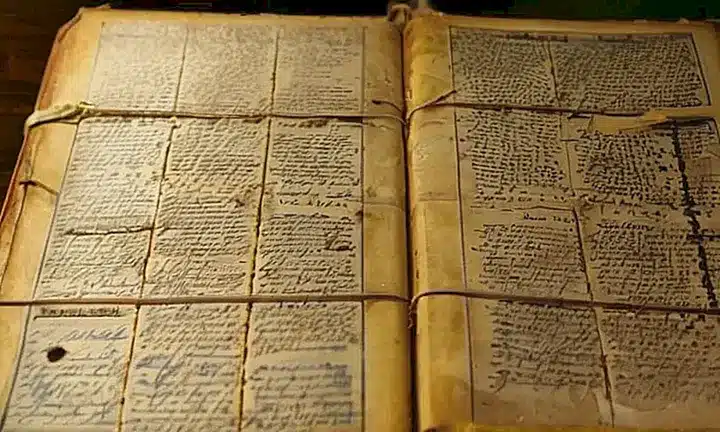 Un viejo cuaderno de cuero desgastado, cubierto por papel de diario, está abierto en una mesa con textos escritos a mano y documentos adjuntos