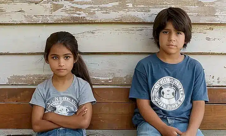 Tres niños de Latin American descent están sentados en una bancal antigua rodeada de paredes coloniales desgastadas, disfrutando del suave luz matinal que entra por grandes ventanas