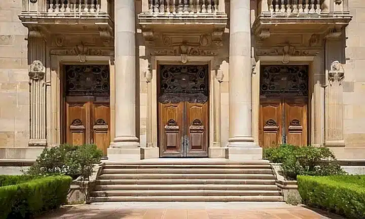 Un antiguo edificio gubernamental español con detalles ornamentales y columnas simétricas se ve desde la calle a través de un paisaje campestre iluminado por el sol