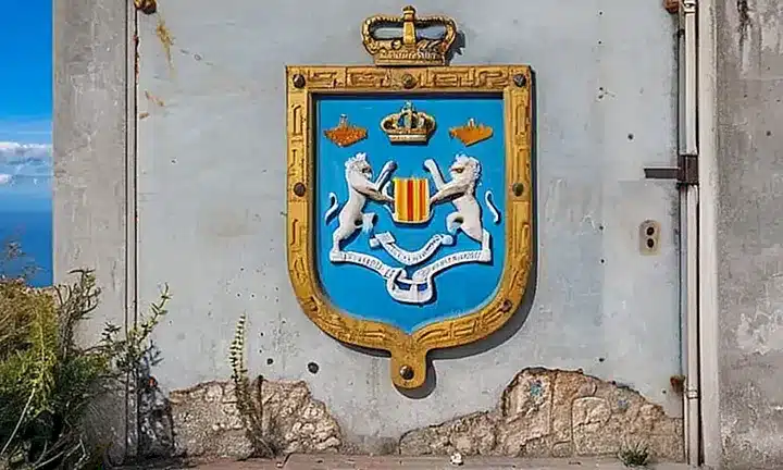 Ante un edificio de gobierno, una pared marrón claro muestra símbolos nacionales desvaídos frente a vegetación mediterránea seca que rodea un portal con cerradura oxidada