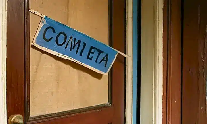 Un pasillo de concreto desgastado y una puerta de madera están junto a un tablero de anuncios sucio y unos ventanales mohosos bajo la luz del sol
