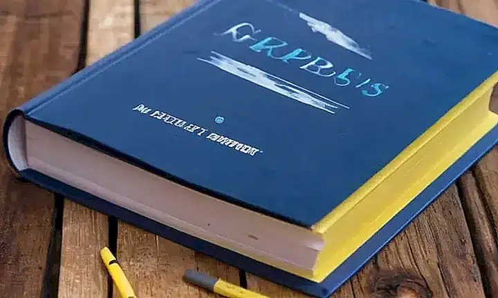 Un libro de papel antiguo descansa en una mesa de madera desgastada acompañado por herramientas de escritura