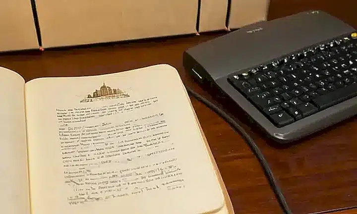 En una mesa desordenada está abierto un libro oscuro con texto negro rodeado de ilustraciones y junto a él hay un ratón que avanza sobre el teclado de una computadora antigua