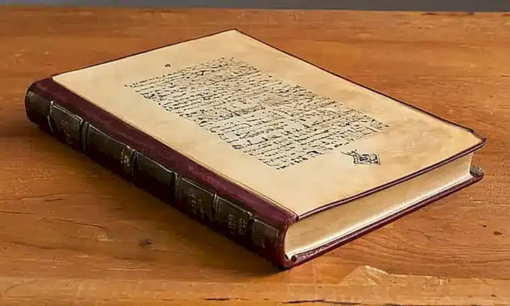 Un libro antiguo de cubierta roja desgastada yace abierto sobre una mesa de madera con notas manuscritas en sus páginas