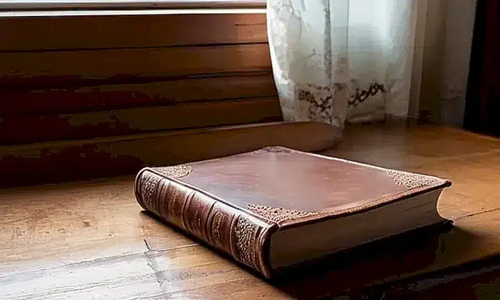 Un libro grande con un largo cojín de cuero marrón oscuro retraído y antiguo reposa en una mesa de madera de roble minimalista con nudos visibles