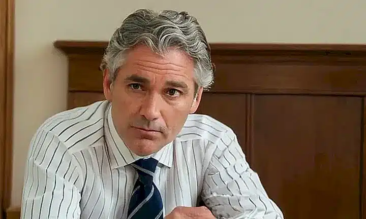 Un hombre de mediana edad con cabello gris ondulado está sentado frente a una mesa de madera en un espacio minimalista, leyendo un libro