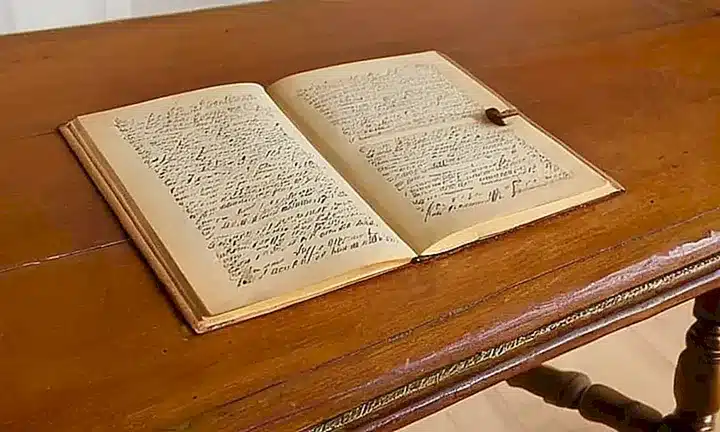 En una habitación antigua, un viejo documento amarillento reposa sobre una mesa de madera, con una pantalla detrás exhibiendo un texto iluminado en español